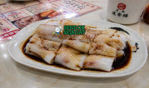 Gulungan chee cheong fun putih halus dengan tekstur licin disiram saus kecap khas, ditaburi wijen dan daun bawang, tampilan mengilap menggugah selera