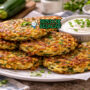 Tumpukan cheesy zucchini fritters Australia autentik dengan permukaan kriuk keemasan, keju cheddar meleleh di dalam, disajikan hangat di piring putih bersama yogurt segar yang menggugah selera