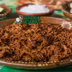 Hidangan chilorio berupa daging babi suwir berwarna merah kecokelatan, terlihat berminyak ringan dengan serpihan cabai, disajikan bersama tortilla dan jeruk nipis