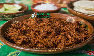 Hidangan chilorio berupa daging babi suwir berwarna merah kecokelatan, terlihat berminyak ringan dengan serpihan cabai, disajikan bersama tortilla dan jeruk nipis
