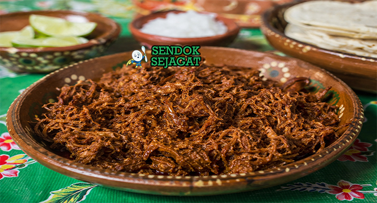Hidangan chilorio berupa daging babi suwir berwarna merah kecokelatan, terlihat berminyak ringan dengan serpihan cabai, disajikan bersama tortilla dan jeruk nipis