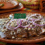 Enmoladas khas Meksiko berupa tortilla jagung berisi ayam suwir yang dilapisi saus mole cokelat gelap kental, dihiasi keju putih dan irisan bawang, tampak lembut dan menggoda