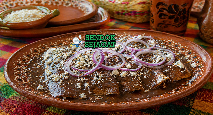 Enmoladas khas Meksiko berupa tortilla jagung berisi ayam suwir yang dilapisi saus mole cokelat gelap kental, dihiasi keju putih dan irisan bawang, tampak lembut dan menggoda