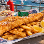 Flathead tail fish & chips khas Australia dengan fillet ikan goreng renyah, kentang tebal kriuk, lemon wedges, dan saus tartar disajikan di kertas koran