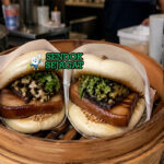 Roti kukus putih lembut terlipat dengan isian daging babi berwarna cokelat karamel, kacang tumbuk, dan daun ketumbar segar