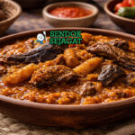 Ikokore khas Nigeria dengan porridge ubi kental berwarna oranye dari palm oil, potongan ikan asap, ponmo, dan daun efirin disajikan di mangkuk