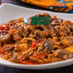 Mangkuk isi ewu khas Nigeria dengan potongan kepala kambing empuk, saus minyak sawit merah kental, taburan bawang bombay dan daun utazi segar