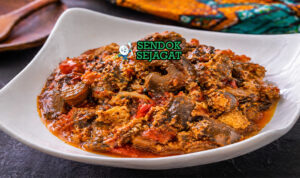 Mangkuk isi ewu khas Nigeria dengan potongan kepala kambing empuk, saus minyak sawit merah kental, taburan bawang bombay dan daun utazi segar