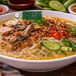 Mangkuk laksa Banjar khas Banjarmasin dengan mie gulungan kenyal, suwiran ikan gabus, kuah santan kekuningan, taburan bawang goreng dan daun bawang di atas meja kayu