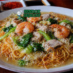 Mie goreng kering khas Makassar dengan tekstur renyah disiram kuah kental berwarna cokelat keemasan, berisi seafood dan sayuran segar