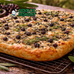 Native pepperberry focaccia disajikan hangat di piring kayu dengan permukaan keemasan berlubang-lubang, taburan sea salt flakes dan bubuk pepperberry yang harum