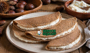 Pancake tipis berwarna cokelat dari tepung kastanye dengan isian ricotta putih lembut di atas piring rustic