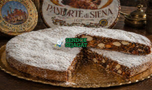 Kue panforte berbentuk bulat pipih dengan permukaan bertabur gula halus, terlihat padat berisi kacang dan buah kering dengan warna cokelat keemasan