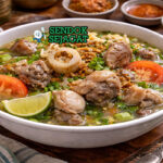 Sop kaki kambing bening khas Betawi dengan kuah jernih, potongan kaki kambing empuk, wortel, kentang, soun, dan rempah disajikan di mangkuk putih dengan bawang goreng tabur