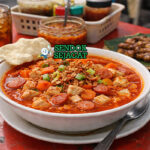 Sop merah khas Surabaya dengan kuah merah cerah, potongan ayam empuk, sosis iris, wortel, dan makaroni disajikan di mangkuk putih dengan bawang goreng tabur