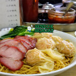 Mi tipis berwarna kuning dengan pangsit udang dalam kuah jernih, dilengkapi sayuran hijau dan topping daging