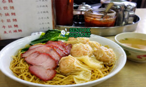 Mi tipis berwarna kuning dengan pangsit udang dalam kuah jernih, dilengkapi sayuran hijau dan topping daging